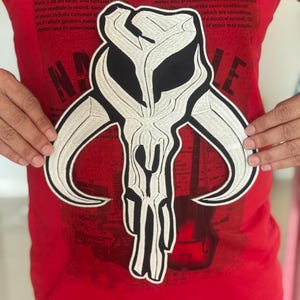Op de afbeelding: Een groot, geborduurd embleem met een gestileerd schedelontwerp met zwarte en witte details. Het embleem wordt tegen een rood t-shirt gehouden met de tekst "MUSIC CITY USA". Het ontwerp heeft een gedurfde, grafische kwaliteit.