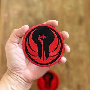 Op de afbeelding: Een rood en zwart geborduurde patch met een symbool dat lijkt op een gestileerde vogel met uitgespreide vleugels. Het symbool bevindt zich op een ronde patch.