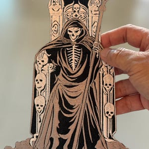 Op de afbeelding: Een geborduurd strijk-patch met een skeletfiguur in een gewatteerde kap, die een zeis vasthoudt en op een troon van schedels zit. De patch is zwart en bruin met witte details.