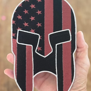 Peut inclure: Un écusson en forme de casque spartiate noir et rouge avec une bordure blanche. Le motif intègre le drapeau américain avec des rayures rouges et des étoiles sur fond noir. L'écusson est tenu dans une main.