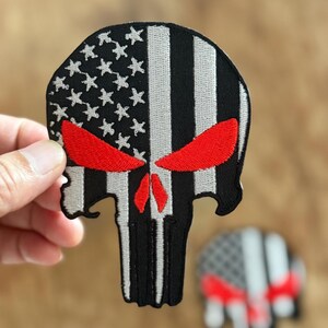 Può includere: Una toppa di teschio con bandiera americana in bianco e nero con occhi rossi. Il teschio ha la forma del logo di Punisher.