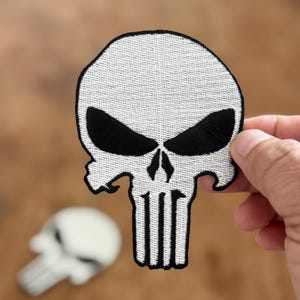 Może przedstawiać: Biała haftowana łatka z czaszką z czarnymi oczami i czarnym obramowaniem. Czaszka ma wyraźną szczękę i szereg pionowych linii w środku.