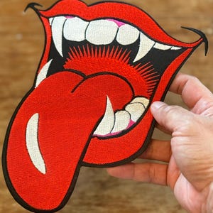 May include: Red embroidered patch of a large, open mouth with sharp teeth and a long, red tongue sticking out. The mouth is outlined in black and the tongue has white details.