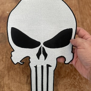 May include: A large white embroidered patch of the Punisher skull. The skull is black with white teeth and a white outline. The skull is on a brown wooden surface.