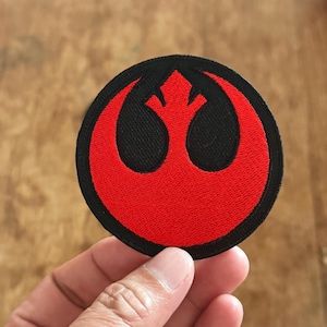 Può includere: Una toppa ricamata rossa e nera con il simbolo dell'Alleanza Ribelle di Star Wars.
