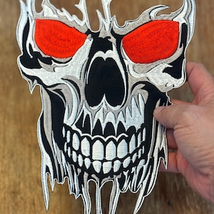 May include: A white skull with red eyes and a dripping effect. The skull is embroidered with white thread on a black background.