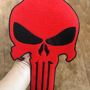 May include: Red embroidered skull patch with black details. The skull has a classic design with a pointed top and a wide jawline. The eyes are black and the teeth are white.