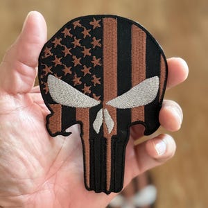Puede incluir: Un parche bordado negro y marrón con el cráneo del Punisher con un diseño de bandera estadounidense. La bandera es marrón y negra con estrellas blancas.