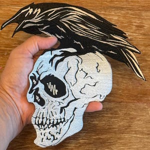 Può includere: Un teschio bianco con un corvo nero appollaiato sopra. Il teschio ha un design dettagliato con una benda nera sull'occhio. Il corvo ha un corpo nero con piume bianche.