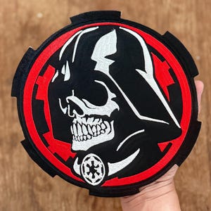 May include: A round, black patch featuring a stylized skull in the likeness of Darth Vader. The design includes white details, a red border, and a symbol at the bottom. The patch is likely for clothing or accessories.