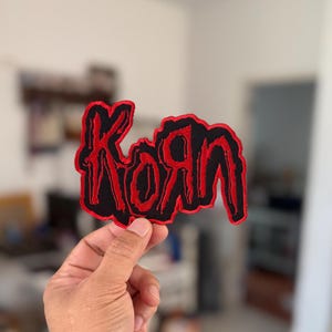 May include: A red and black embroidered patch with the word "Korn" in a stylised font.