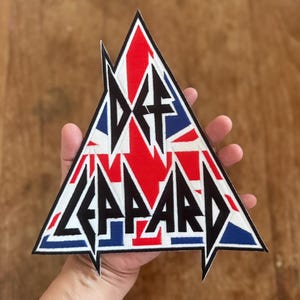 May include: A triangular shaped iron-on patch with the band name "Def Leppard" in black lettering and a Union Jack flag design in red, white, and blue.