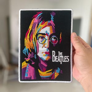 Könnte beinhalten: Gestickter Aufnäher mit einem farbenfrohen, abstrakten Porträt von John Lennon und dem Text "The Beatles" in Weiß.