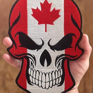 May include: An embroidered patch featuring a skull with a Canadian flag design. The skull is red, white and black, with a menacing expression and a large, toothy grin. The Canadian flag is on the forehead of the skull, with a red maple leaf on a white background.