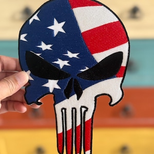 Puede incluir: Un diseño de bandera americana roja, blanca y azul en forma de calavera. La calavera tiene un contorno negro y ojos negros.