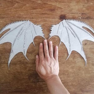 May include: Two light grey, textured dragon wing cutouts featuring a jagged, pointed edge design. Each wing displays a detailed, ribbed structure with sharp, protruding spikes along the top and side edges, crafted from a flat, firm material.