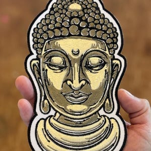 Puede incluir: Parche bordado con la cabeza de un Buda dorado con los ojos cerrados y una expresión serena. El parche tiene un borde blanco y un contorno negro. El diseño se sostiene en una mano.