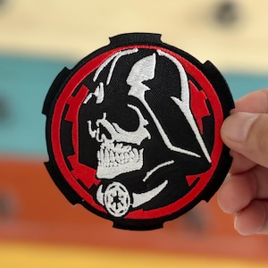 May include: A black and white embroidered patch featuring a skull wearing a Darth Vader helmet. The skull is encircled by a red circle with a white symbol at its centre.