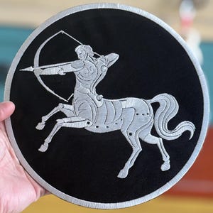 Puede incluir: Un parche bordado en plata sobre un fondo negro que representa un centauro con arco y flecha. El centaur está mirando hacia la derecha y tiene un torso humano y un cuerpo de caballo.