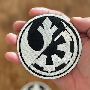 Pode incluir: Um remendo bordado em preto e branco com o símbolo da Aliança Rebelde e o símbolo imperial, dividido ao meio.