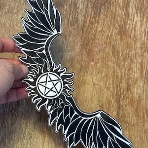 Peut inclure: Patch brodé noir et blanc représentant une paire d'ailes d'ange avec un pentagramme à l'intérieur d'un soleil éclatant.