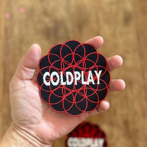 May include: A black and red embroidered patch with the word "COLDPLAY" in white lettering. The patch features a circular design with multiple overlapping circles.
