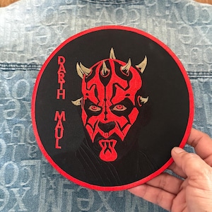 May include: A black and red round embroidered patch with a red Darth Maul face and the text "Darth Maul" in gold lettering.