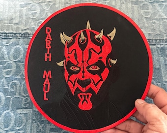 Può includere: Una toppa rotonda ricamata in nero e rosso con un viso rosso di Darth Maul e il testo "Darth Maul" in lettere dorate.