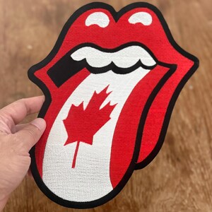 May include: A red and white tongue and lips patch with a Canadian flag design. The tongue is sticking out and the lips are closed. The patch is made of embroidered fabric.