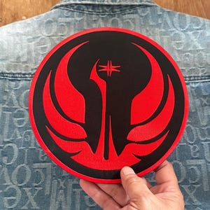 May include: A round, black patch with a red emblem featuring a stylized bird or wing design. The patch is being held up against a denim jacket. The emblem has a central vertical element with a cross-like shape at the top.