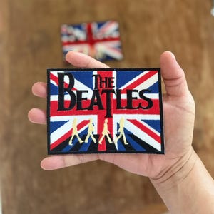 Könnte beinhalten: Ein gestickter Aufnäher mit der Union Jack Flagge, den Worten "The Beatles" und einer Silhouette der Bandmitglieder, die über einen Zebrastreifen gehen.