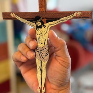 May include: Embroidered patch of Jesus Christ on a brown cross. The patch is detailed with a crown of thorns, a white loincloth, and red nail marks.