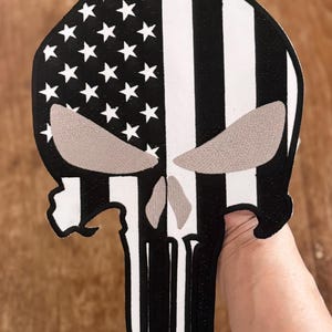 Könnte beinhalten: Schwarz-weiß gestickter Patch des Punisher-Schädels mit amerikanischem Flaggen-Design. Der Schädel ist weiß umrandet und die Flagge ist in der Mitte geteilt, mit den Sternen links und den Streifen rechts.