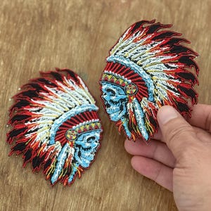 Puede incluir: Dos parches bordados para planchar que representan una calavera con un tocado de nativo americano con plumas rojas, blancas y azules.