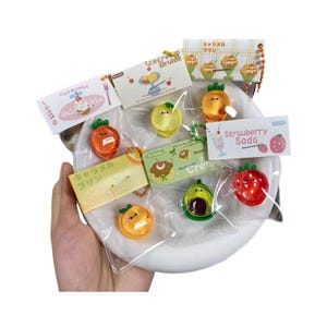 May include: A collection of small, colourful food-themed charms with smiling faces, each individually packaged in clear plastic with a decorative paper header. The charms include designs like strawberry, avocado, orange, and pudding, with text labels.