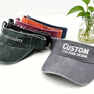 Könnte beinhalten: Eine Sammlung von Sonnenblenden in verschiedenen Farben, darunter Grau, Grün, Orange und Blau. Die graue Sonnenblende im Vordergrund trägt den weißen Schriftzug "CUSTOM YOUR DESIGN". Die grüne Sonnenblende trägt den weißen Schriftzug "Embroidery".