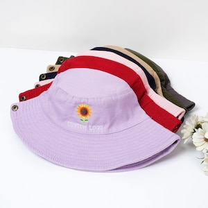 May include: A stack of colorful bucket hats in various colors, including red, pink, and lavender. The lavender hat features an embroidered sunflower and the text "CUSTOM LOGO". The hats are made of a soft fabric and have a classic design.