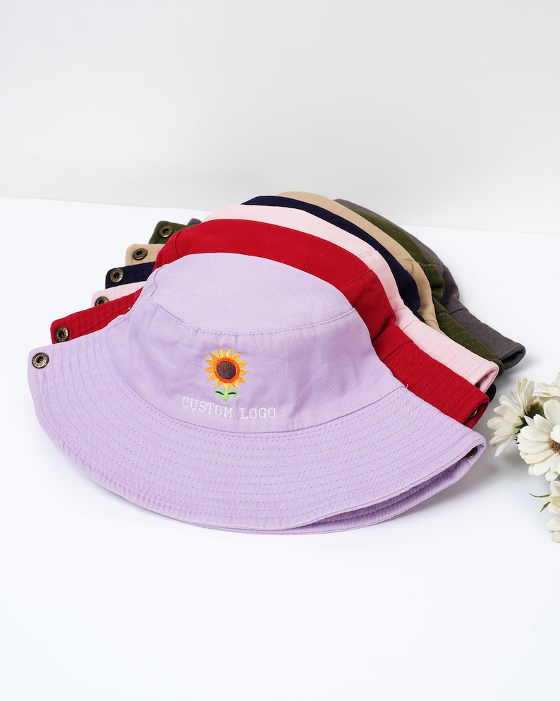 May include: A stack of colorful bucket hats in various colors, including red, pink, and lavender. The lavender hat features an embroidered sunflower and the text "CUSTOM LOGO". The hats are made of a soft fabric and have a classic design.