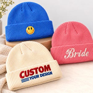 May include: Three ribbed knit beanies in blue, pink, and cream. The blue beanie features a yellow smiley face patch. The pink beanie has the word Bride embroidered in white. The cream beanie displays the text CUSTOM YOUR DESIGN in red and blue.