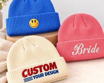 May include: Three ribbed knit beanies in blue, pink, and cream. The blue beanie features a yellow smiley face patch. The pink beanie has the word Bride embroidered in white. The cream beanie displays the text CUSTOM YOUR DESIGN in red and blue.