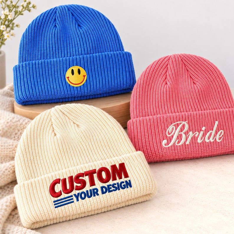 May include: Three ribbed knit beanies in blue, pink, and cream. The blue beanie features a yellow smiley face patch. The pink beanie has the word Bride embroidered in white. The cream beanie displays the text CUSTOM YOUR DESIGN in red and blue.