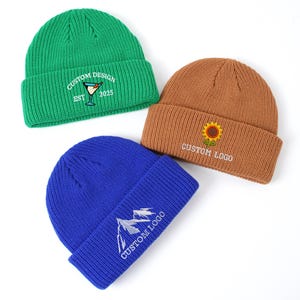May include: Three ribbed knit beanies in green, brown, and blue. The green beanie features a martini glass graphic with the text "CUSTOM DESIGN EST 2025". The brown beanie has a sunflower graphic and "CUSTOM LOGO". The blue beanie shows a mountain graphic and "CUSTOM LOGO".