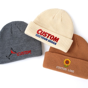 May include: Three ribbed knit beanies in charcoal gray, cream, and tan. The gray beanie features a martini glass and the word CUSTOM. The cream beanie displays CUSTOM YOUR DESIGN in red and blue. The tan beanie shows a sunflower and CUSTOM LOGO.