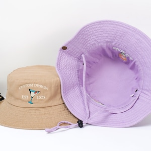 May include: Two bucket hats. One is tan with a cocktail glass design and the text "CUSTOM DESIGN EST 2025". The other is purple, with a drawstring and a floral design inside. Both hats are made of a soft fabric.