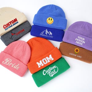 May include: A collection of nine ribbed knit beanies in various colors including cream, gray, blue, purple, brown, pink, orange, and green. Each beanie features embroidered text or graphics such as Custom Your Design, Bride, Mom, and Custom Text.