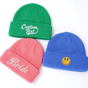May include: Three knit beanies in green, pink, and blue. The green beanie features white embroidered text that reads Custom Text. The pink beanie has white cursive embroidery reading Bride. The blue beanie features a yellow smiley face patch.