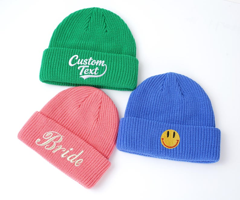 May include: Three knit beanies in green, pink, and blue. The green beanie features white embroidered text that reads Custom Text. The pink beanie has white cursive embroidery reading Bride. The blue beanie features a yellow smiley face patch.