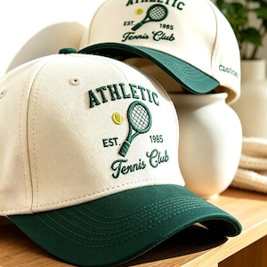 Pode incluir: Dois bonés de beisebol em tom branco sujo com aba verde-escura. A frente apresenta um bordado verde-escuro com o texto ATHLETIC Tennis Club, uma raquete de ténis verde e uma pequena bola de ténis amarela. Um boné exibe EST. 1985, o outro EST. 1955.