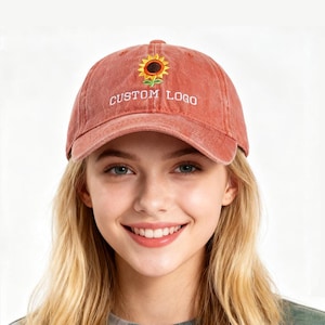 Può includere: Un cappellino da baseball rosa sbiadito con un ricamo di girasole e la scritta "CUSTOM LOGO". Il cappellino è indossato da una persona con i capelli biondi. Un accessorio casual.