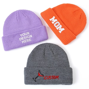May include: Three knit beanies in lavender, orange, and dark gray. The lavender beanie features white text reading YOUR DESIGN HERE. The orange beanie has white text reading MOM. The dark gray beanie displays a red cocktail glass icon and the red text CUSTOM.